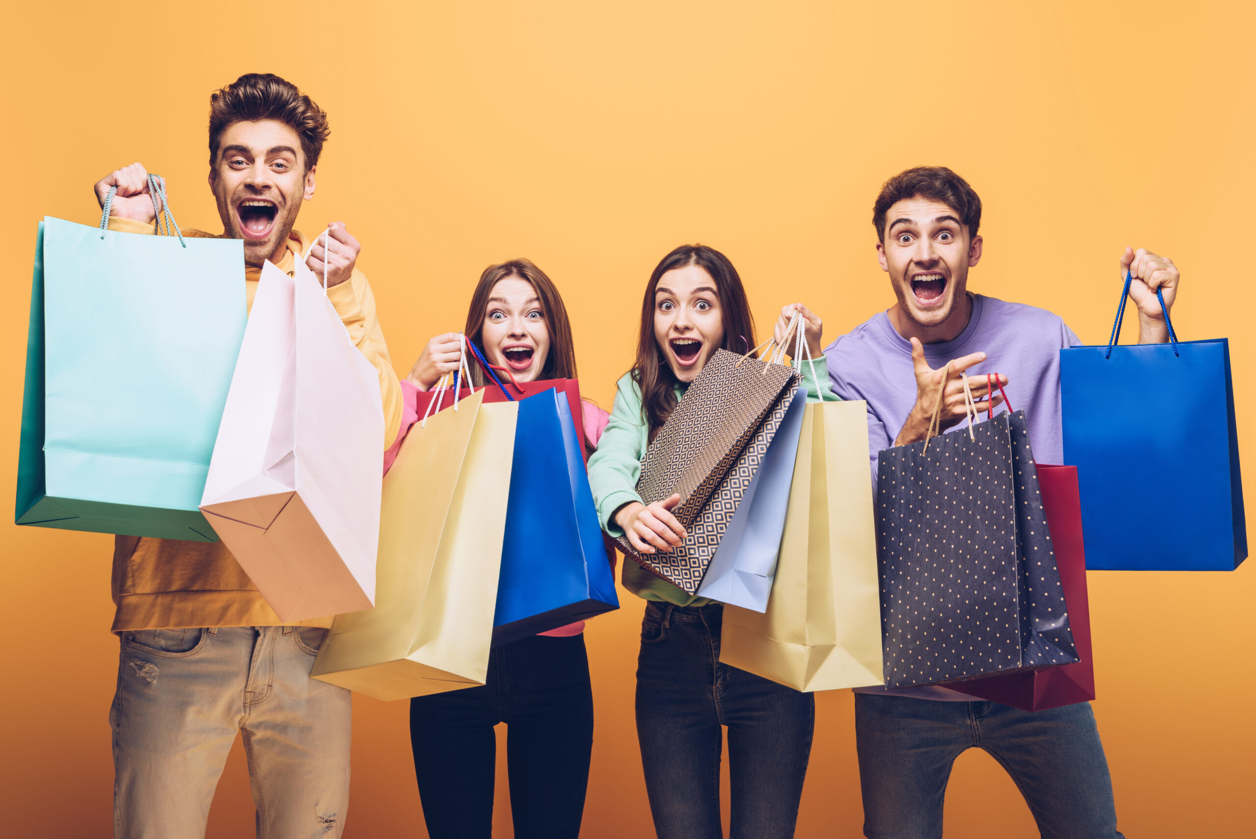 excited young friends shouting and holding shopping bags together, isolated on yellow